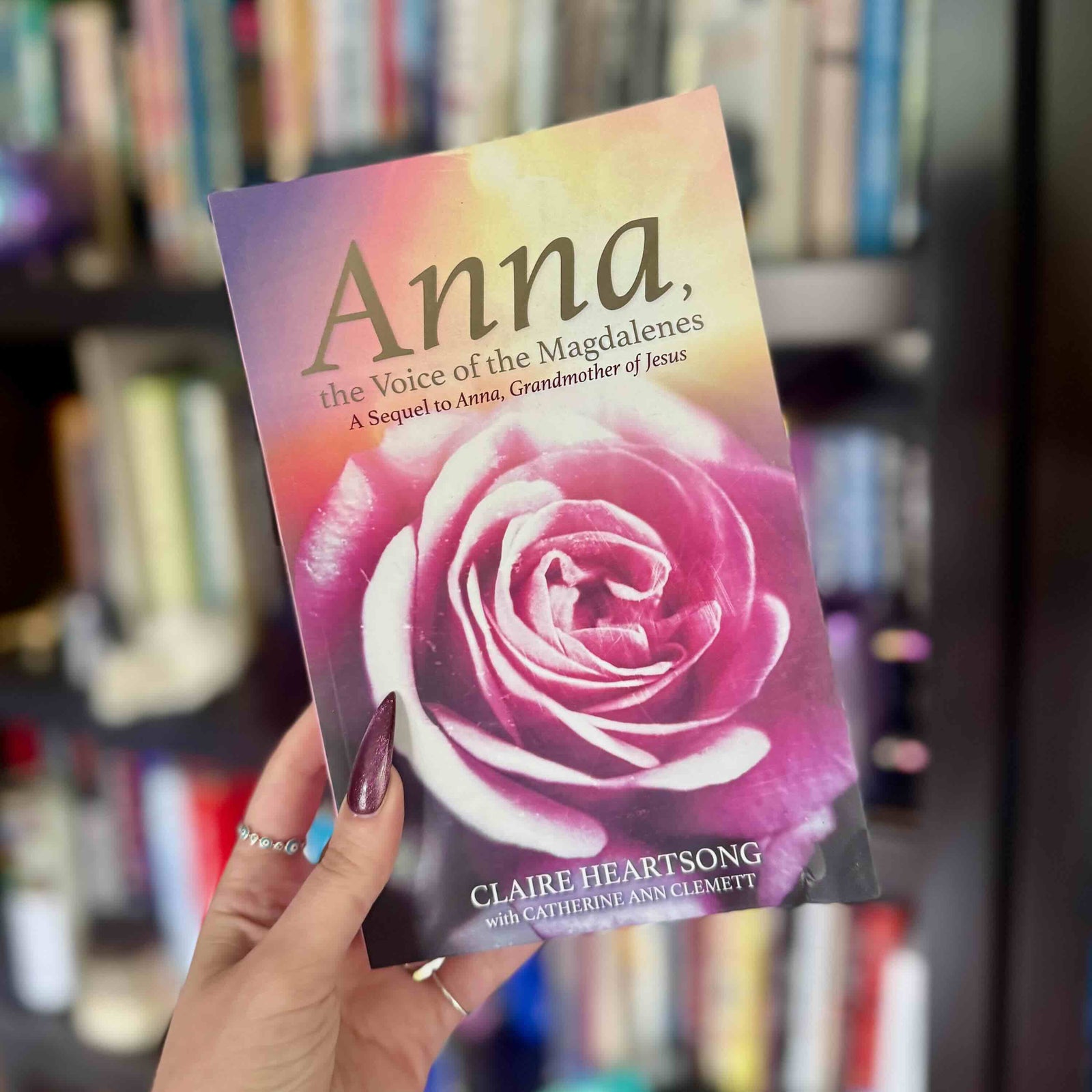 Book titled 'Anna, the Voice of the Magdalenes'  by Claire Heartsong held in front of a bookshelf, featuring a cover photo of a pink rose.