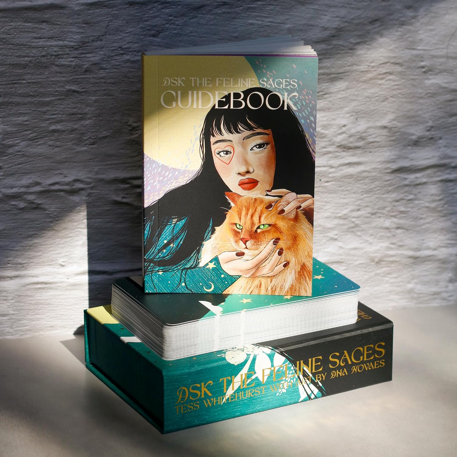 A boxed card deck titled "Ask the Feline Sages" in gold text with artwork of a gray cat resting on a woman's shoulders, with teal, black, and cream.
