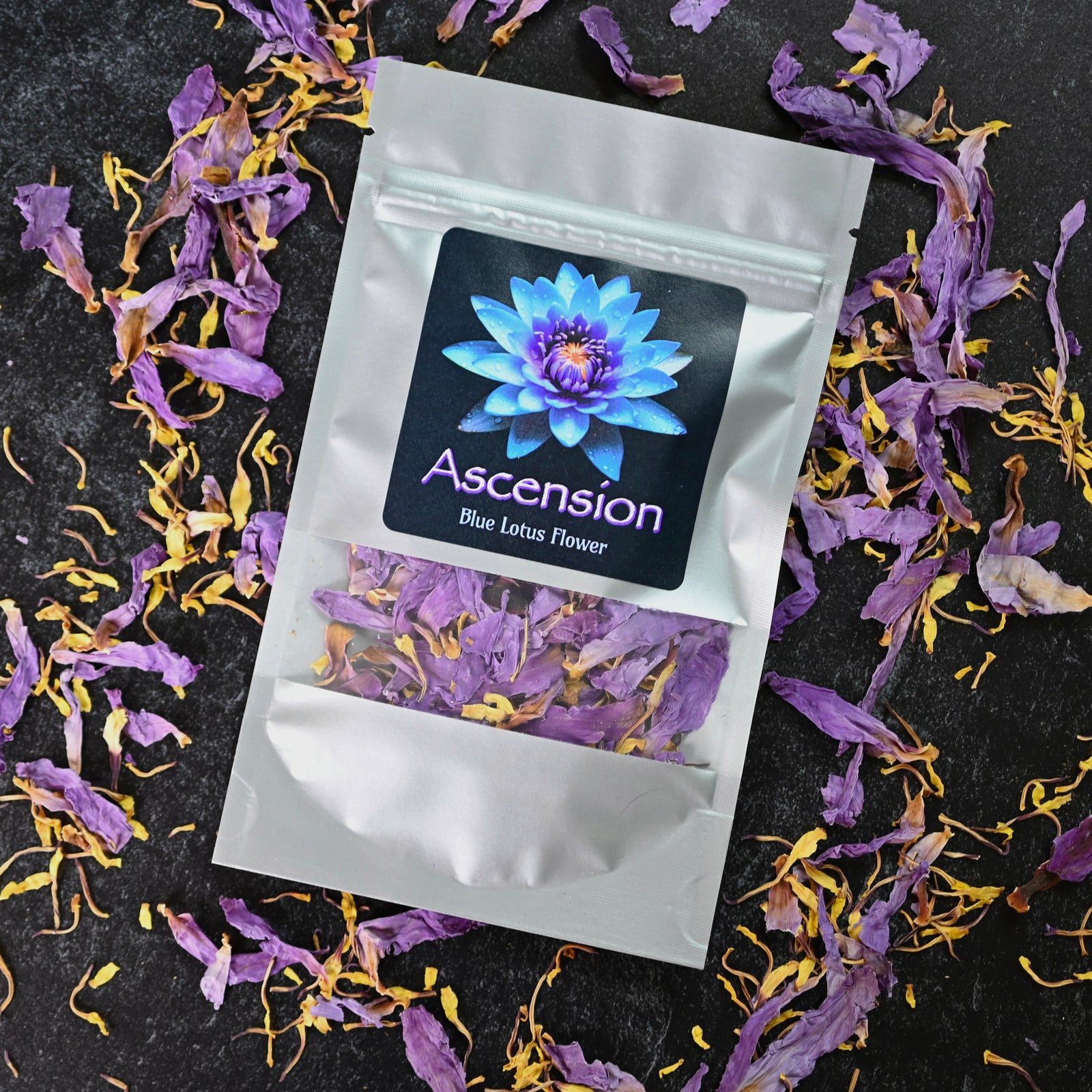 Two silver pouches of Ascension Blue Lotus Flower, with visible dried petals and a vibrant lotus label, surrounded by scattered petals on black background.