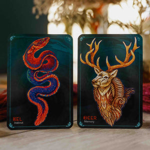 Two oracle cards on wood surface, with a blurred background. Left card shows a red and blue eel, right card shows golden brown deer on dark backgrounds.
