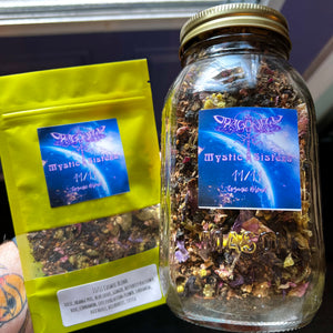 A tattooed hand holds a yellow packet of 11/11 Cosmic Blend Tea next to a glass mason jar filled with similar dried herbs. Both containers have labels reading "Mystic Sisters 11/11 Cosmic Blend" with colorful abstract designs. The unique blend exudes cosmic vibes, set against a blurred window background.