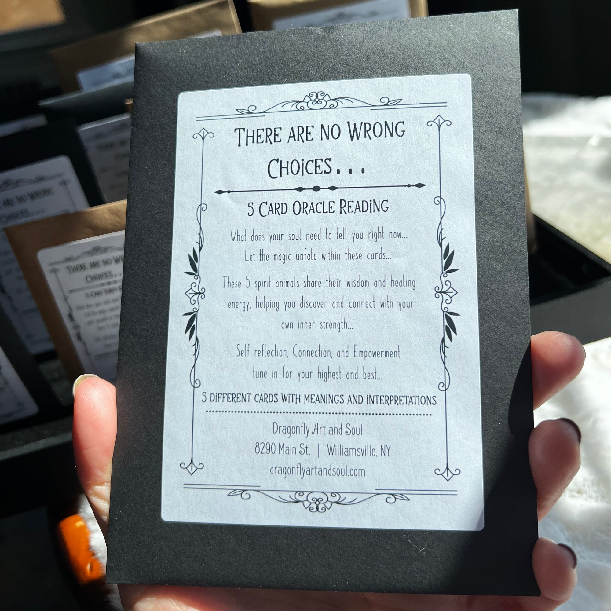 A hand holding a black card with a white label titled "5 Card Mini Oracle Reading." The text describes a reading service that guides your soul's journey through self-reflection and empowerment, offering insight. Located at 8290 Main St., Williamsville, NY.