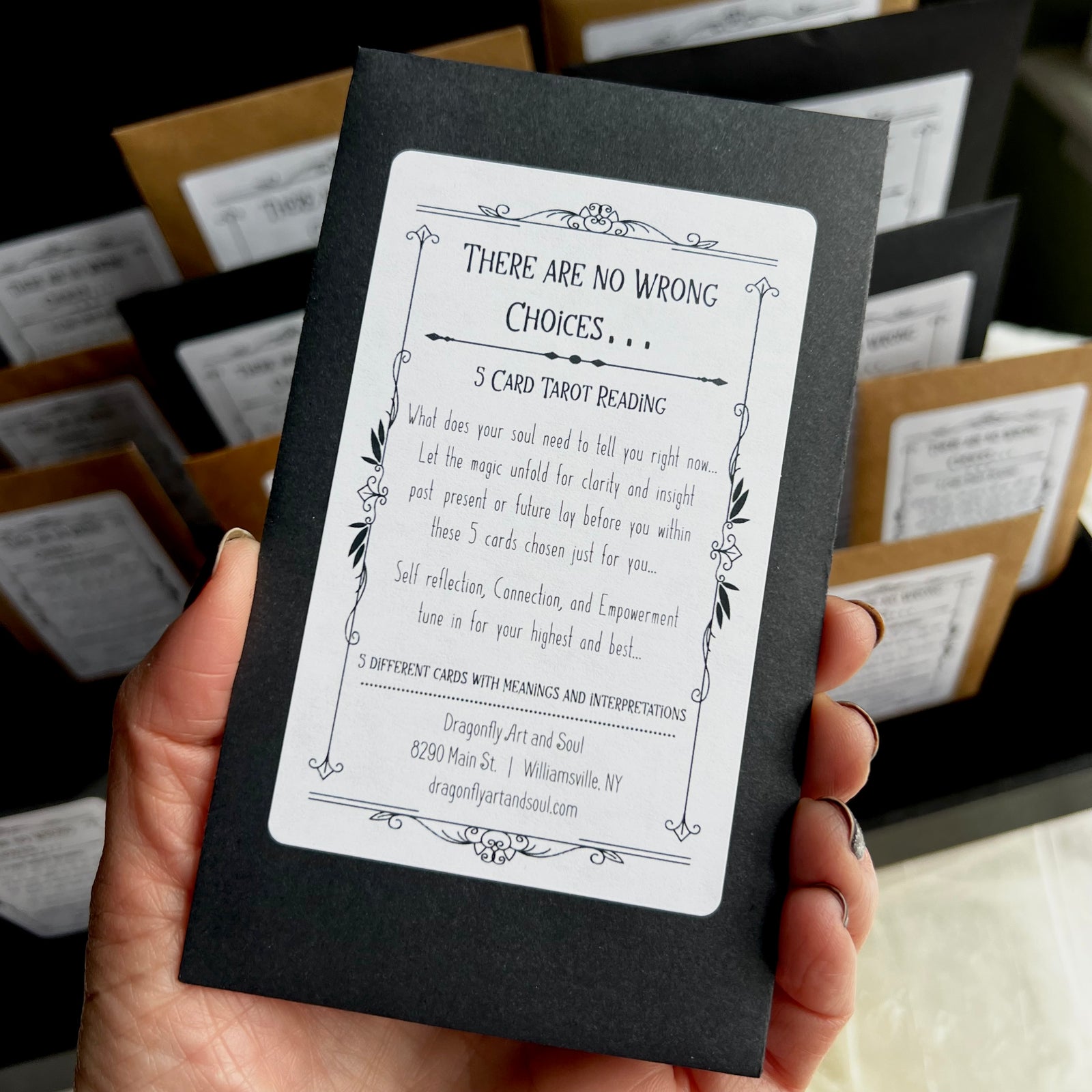 A hand holds a black envelope with a label that reads "There Are No Wrong Choices... 5 Card Mini Tarot Reading." This reading, provided by Dragonfly Art and Soul in Williamsville, NY, offers self-reflection, connection, and empowerment.