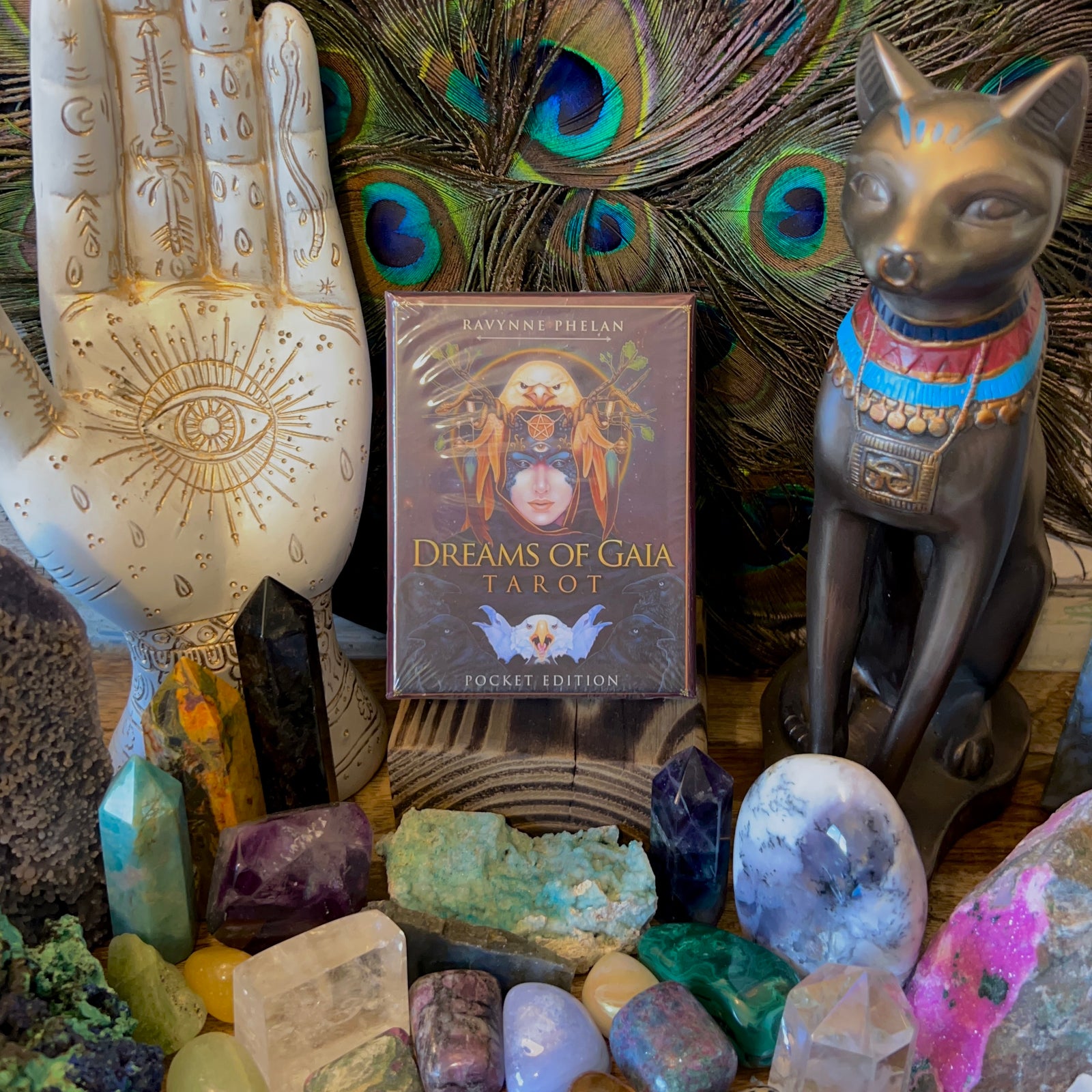A collection of crystals and stones surrounds a "Dreams of Gaia Tarot Pocket Edition" deck, with a peacock feather backdrop. A decorative hand sculpture and an Egyptian cat figure add to the mystical display, fostering intuitive growth and connection to one's divine self.