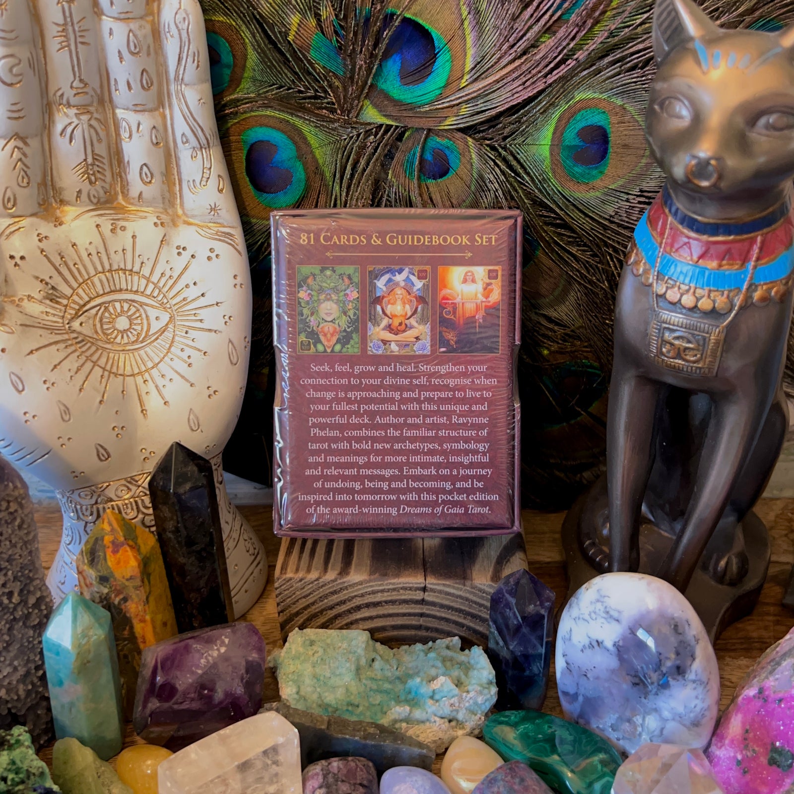 A collection of crystals and stones surrounds a "Dreams of Gaia Tarot Pocket Edition" deck, with a peacock feather backdrop. A decorative hand sculpture and an Egyptian cat figure add to the mystical display, fostering intuitive growth and connection to one's divine self.