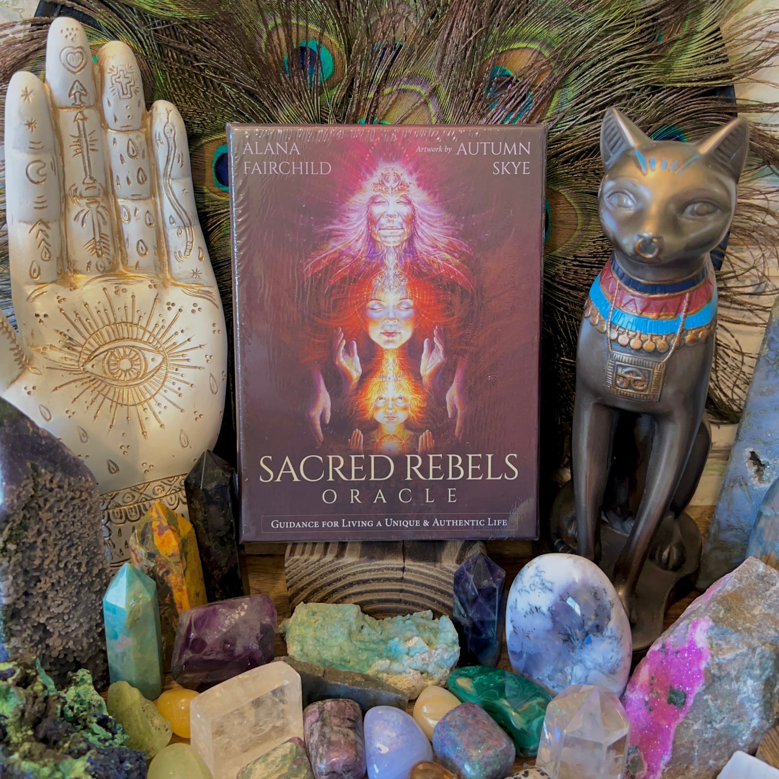 A collection of items including a deck of cards titled 'Sacred Rebels Oracle', surrounded by various crystals, a peacock feather and more