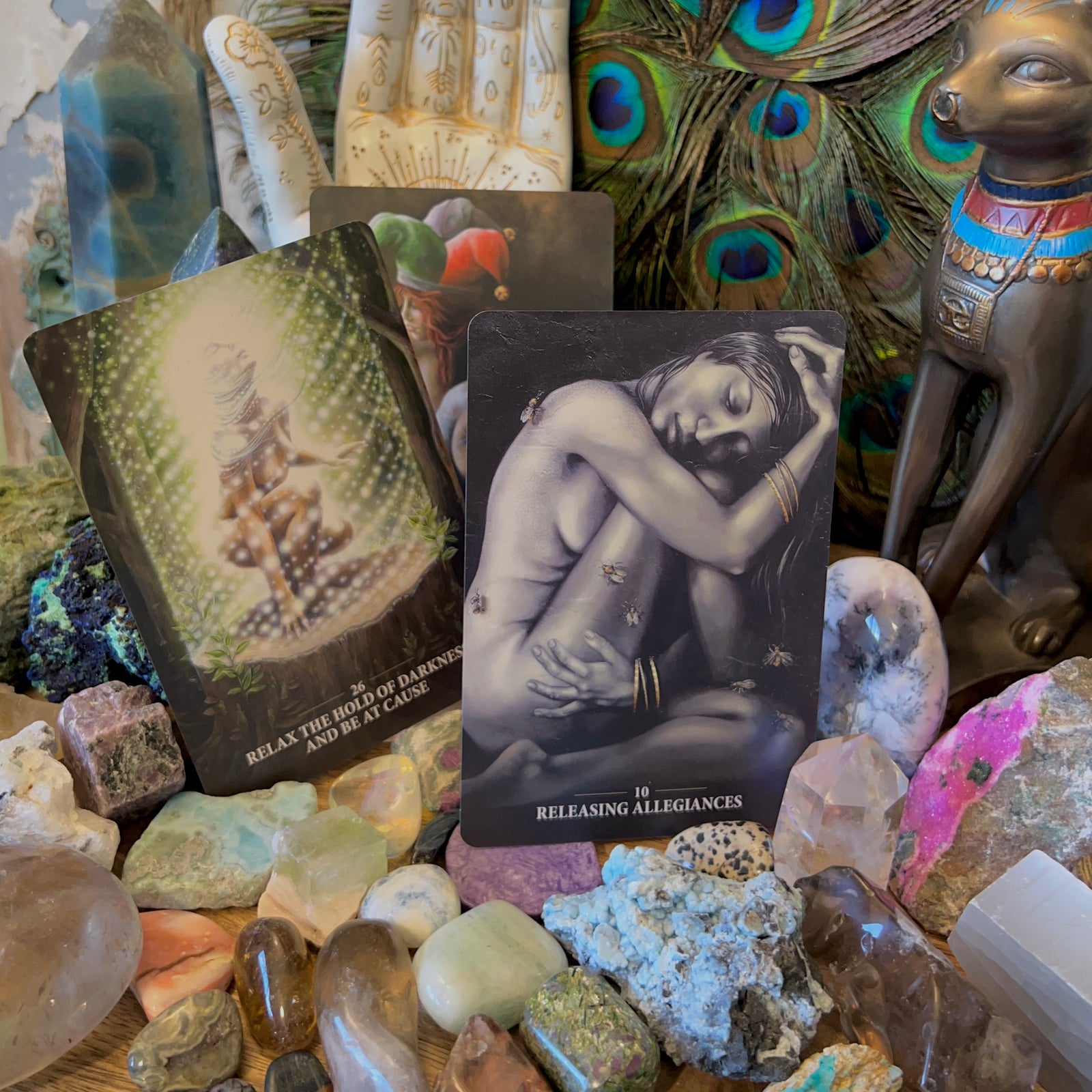 A collection of items including a deck of cards titled 'Sacred Rebels Oracle', surrounded by various crystals, a peacock feather and more