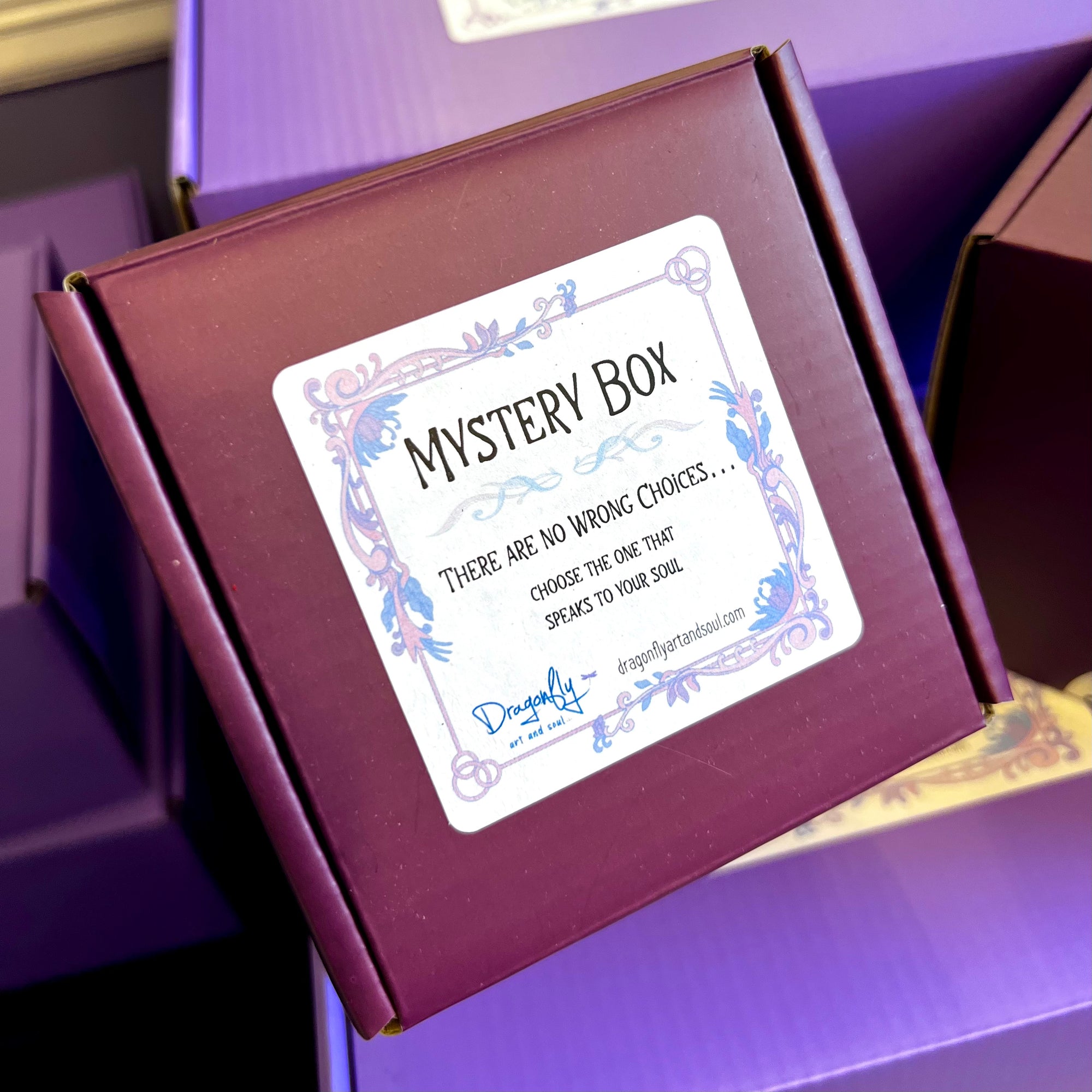 A purple Mystery Box labeled with a decorative design around the text. The label reads: "There are no wrong choices... Choose the one that speaks to your soul." Several other purple Mystery Boxes, each potentially hiding crystals or candles, are visible in the background.