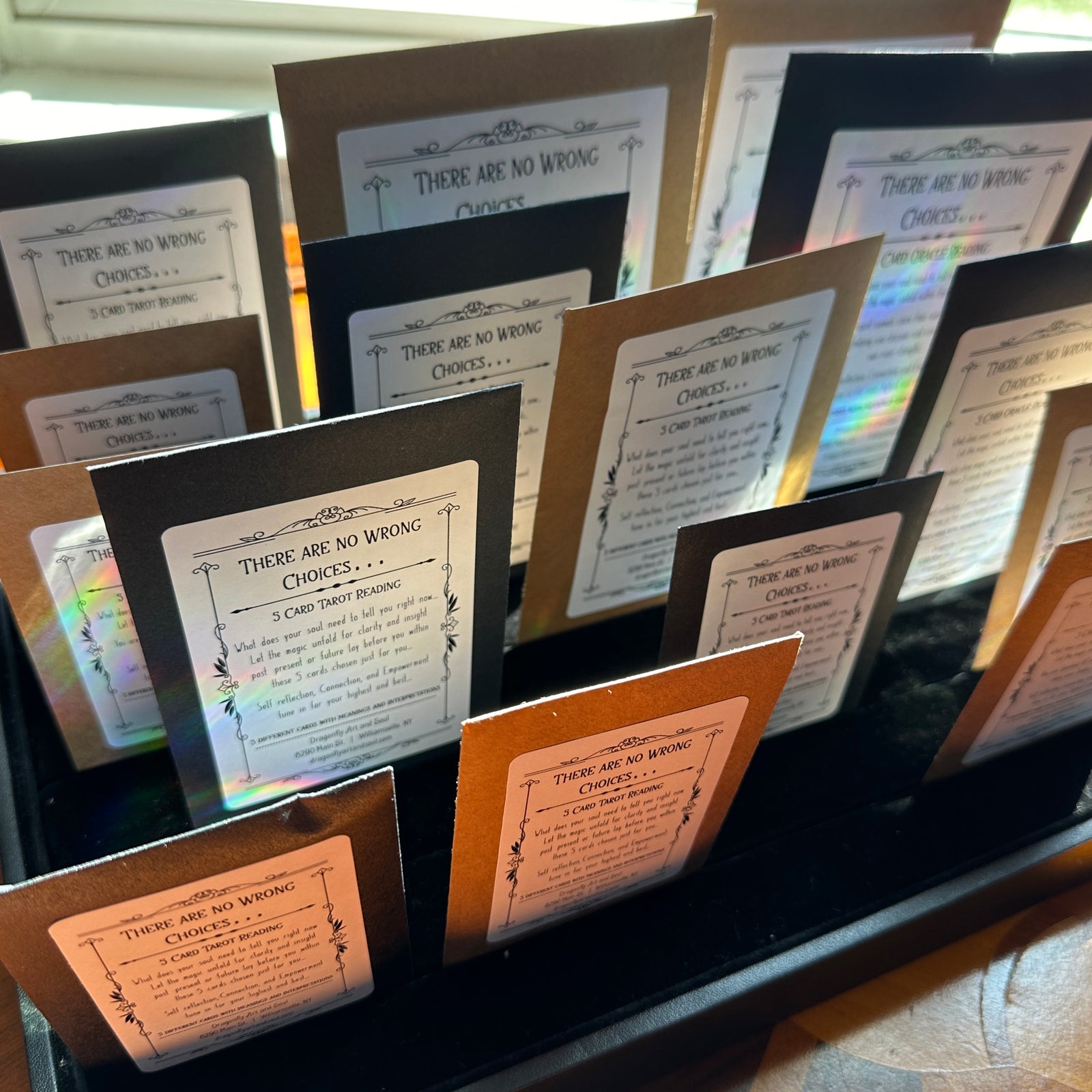 A hand holds a black envelope with a label that reads "There Are No Wrong Choices... 5 Card Mini Tarot Reading." This reading, provided by Dragonfly Art and Soul in Williamsville, NY, offers self-reflection, connection, and empowerment.