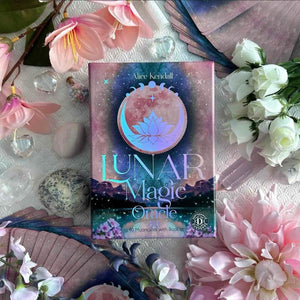 oracle deck titled 'Lunar Magic Oracle' surrounded by flowers and crystals on a soft surface
