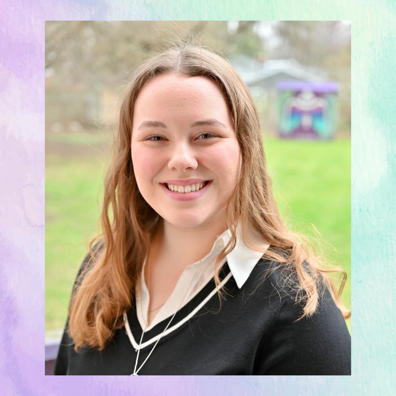 Person with long blonde hair wearing a black sweater with a white collar outdoors.