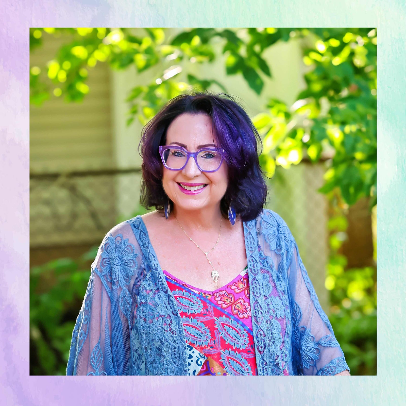Woman with purple hair and glasses standing outdoors with greenery in the background