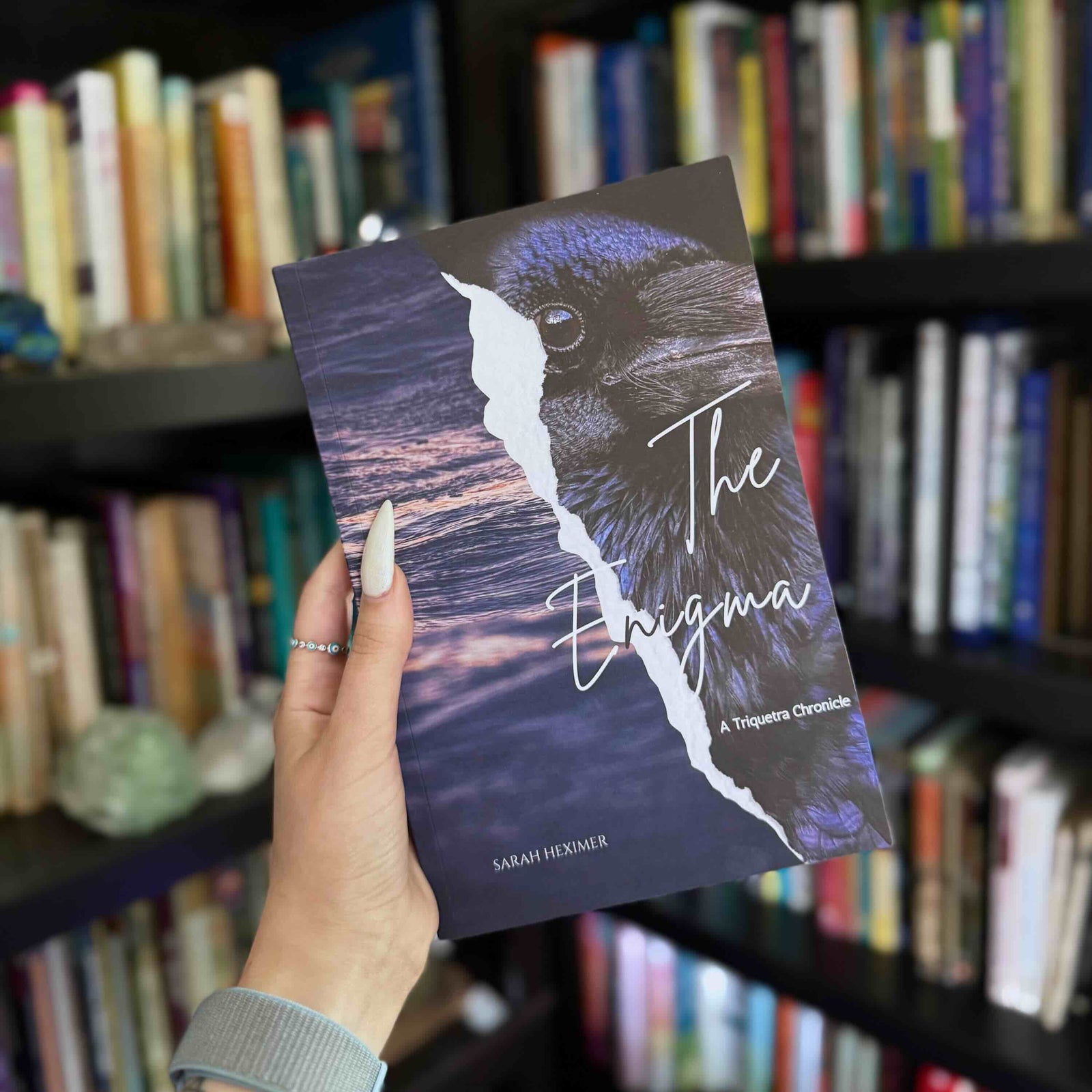 A hand holds a book titled "The Enigma: A Triquetra Chronicle" by Sarah Heximer, features a dark raven with a torn-paper effect revealing water.