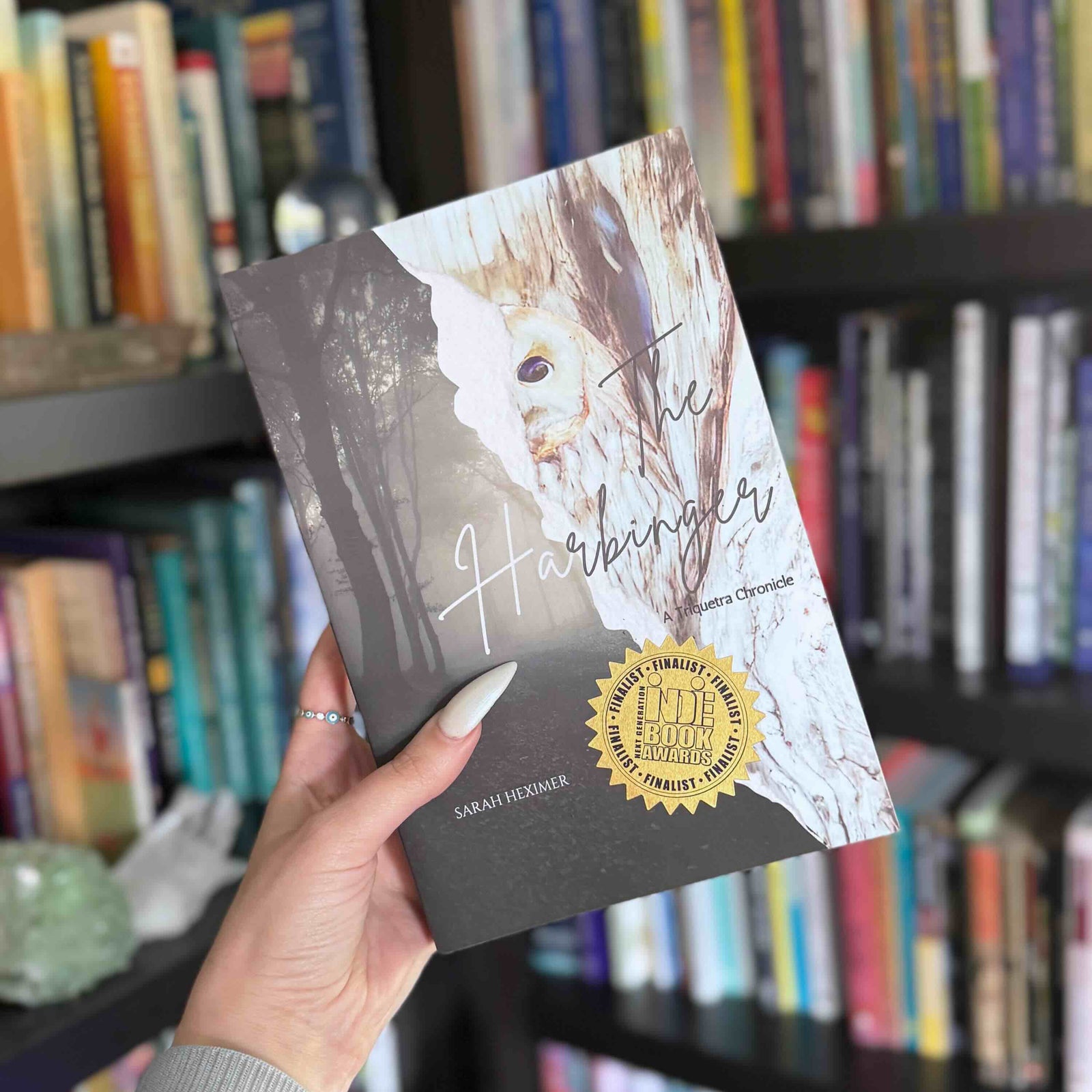 A hand holds a book titled 'The Harbinger' by Sarah Heximer, showing the front cover with a gold seal, against a blurred background of bookshelves.