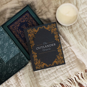 Oracle cards titled 'The Outlander Oracle' on a textured surface with a candle.