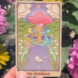 Card titled 'The Crossroad' with a colorful illustration of a tree, flowers, and a cat, held against a floral background.