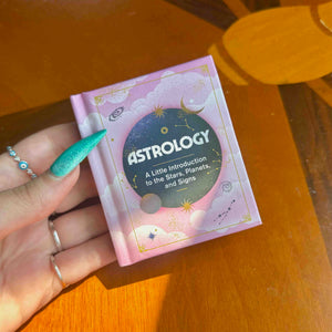 A hand holds a mini pink book titled 'ASTROLOGY: A Little Introduction to the Stars, Planets, and Signs' on a warm wooden table.