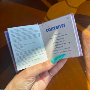 A hand holds a mini open book showing the 'CONTENTS' page and copyright text. The book is photographed over a warm brown wooden surface.