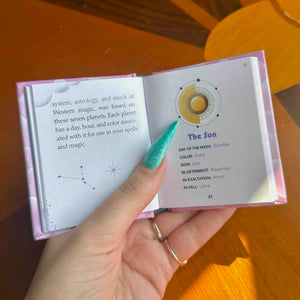 A hand holds an open mini book showing printed pages about the Sun, astrology, and Western magic, photographed on a warm wooden table.