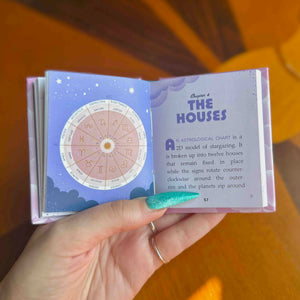A hand holds a mini hardcover book showing an open astrology chart page titled "Chapter 1: THE HOUSES".