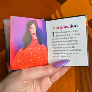 Hand holding a small book with an illustration of a woman and text on a wooden surface