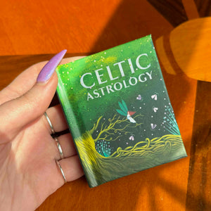 Hand holding a small green book labeled 'Celtic Astrology' on a wooden surface