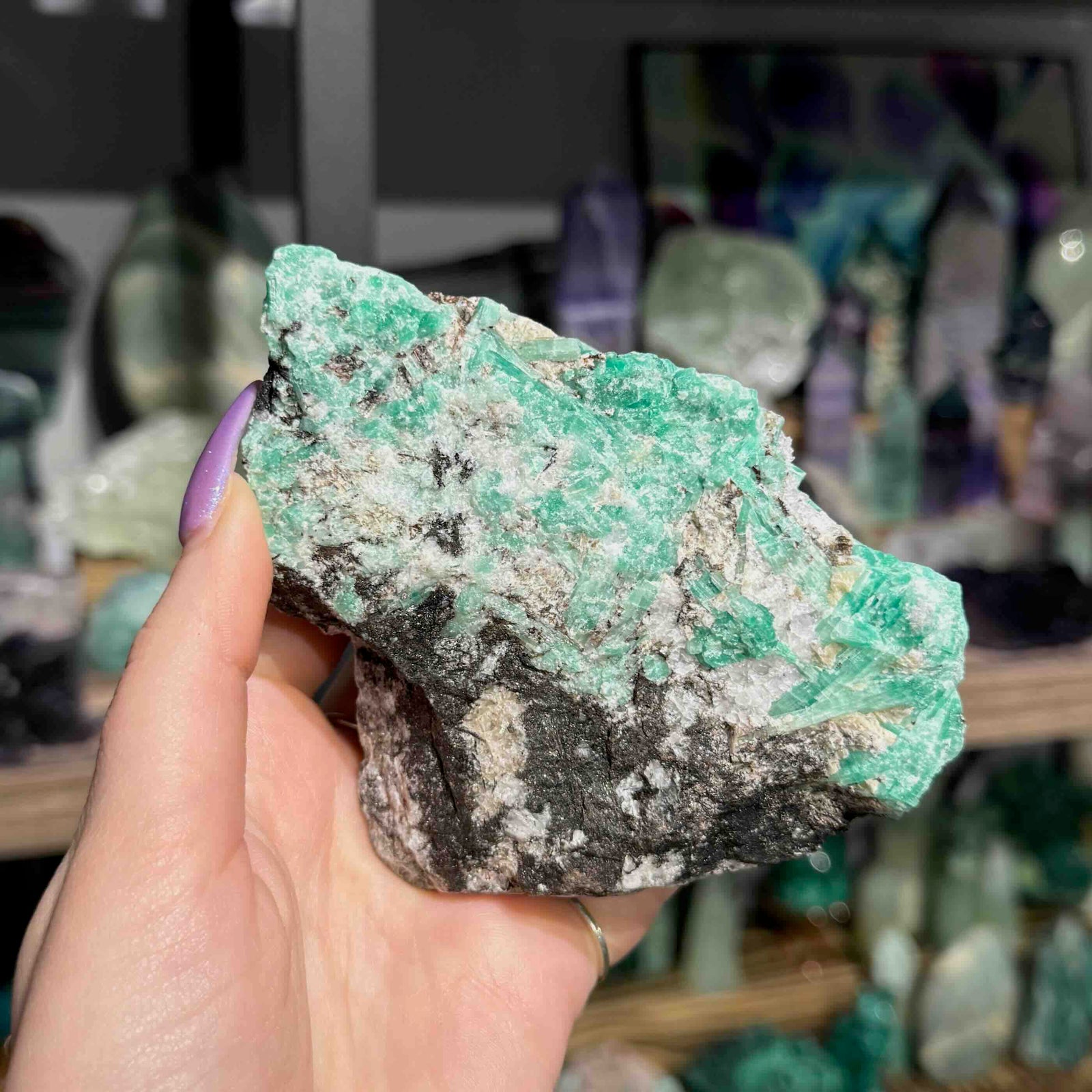Hand holding a large green and black mineral crystal with a blurred background of more crystals.