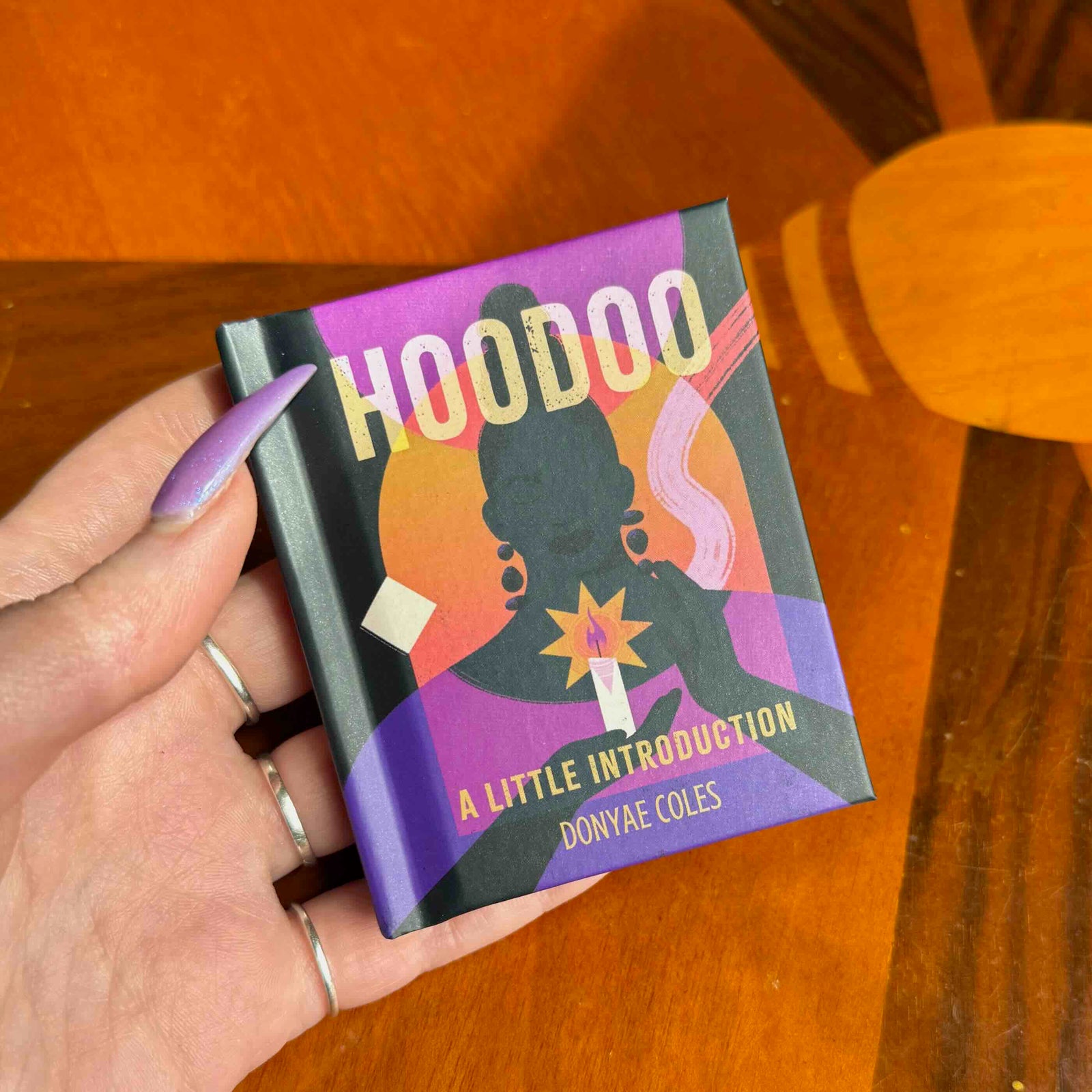 Hand holding a book titled 'Hoodoo: A Little Introduction' by Donyae Coles on a wooden surface.