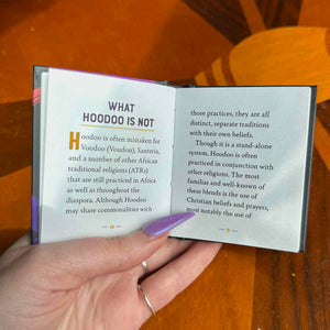 Hand holding an open book with text about Hoodoo, on a wooden surface.