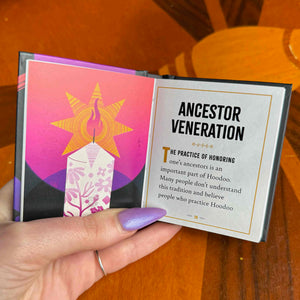 small book opened with 'Ancestor Veneration' text and colorful design held by a hand on a wooden surface