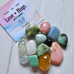 Colorful stones on a wooden surface with a card reading 'Love + Hugs' in the background.