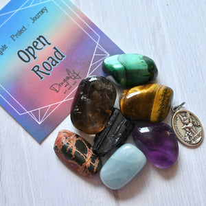 Colorful tumbled stones and a small metal pendant on a white surface with a 'Open Road' card in the background.