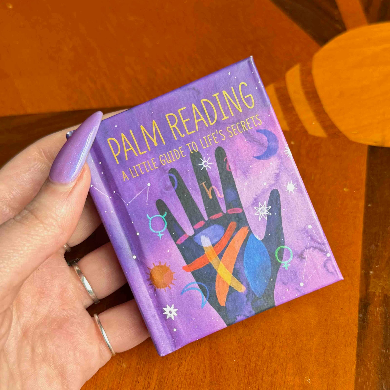 Hand holding a small book titled 'Palm Reading: A Little Guide to Life's Secrets' on a wooden surface.