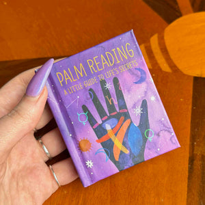 Hand holding a small book titled 'Palm Reading: A Little Guide to Life's Secrets' on a wooden surface.