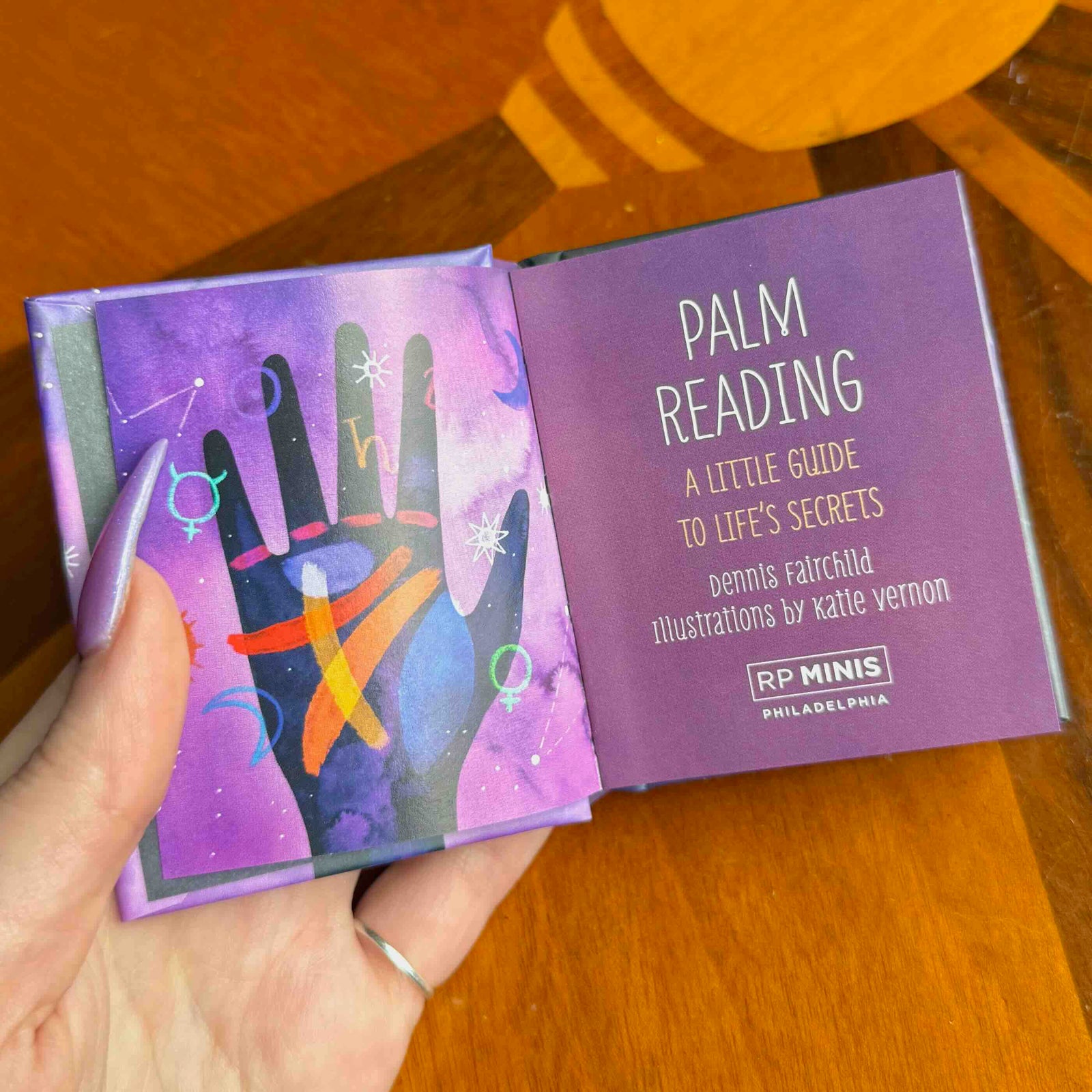 Hand holding a small book titled 'Palm Reading: A Little Guide to Life's Secrets' on a wooden surface.