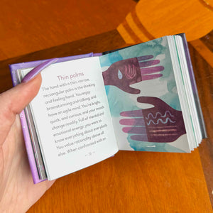 Hand holding a small open book with illustrations of hands on a wooden surface