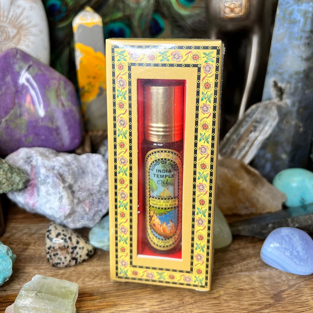 A bottle of India Temple Oil is displayed in its decorative packaging. The box is yellow with ornate designs and a clear window revealing the bottle inside, evoking the meditation scent of holy temples in India. Surrounding the box are various colorful crystals and stones, placed on a wooden surface.