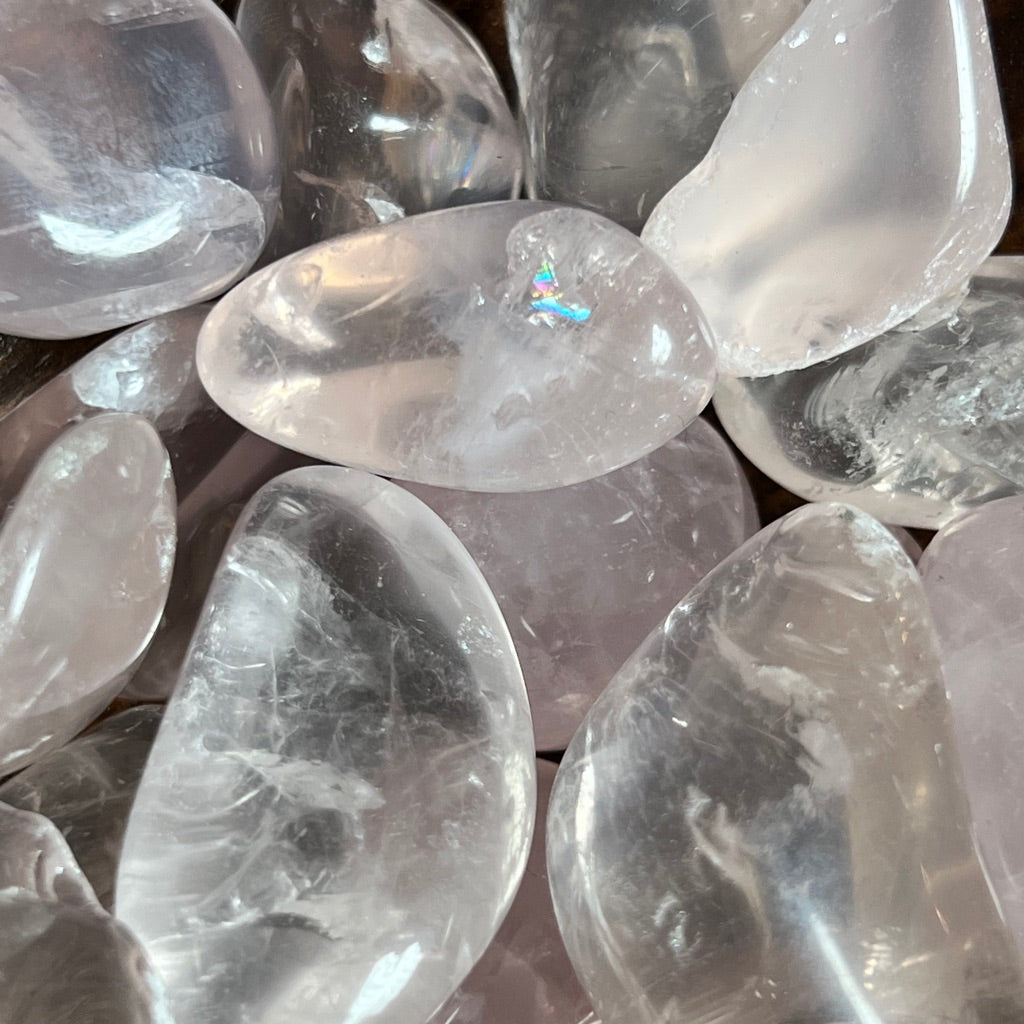 A wooden bowl filled with polished Rose Quartz [high grade] stones sits on a textured wooden surface. The stones vary in size and have a translucent, pink hue, known for their connection to the heart chakra. The bowl's natural wood grain complements the smooth, glossy appearance of the quartz.