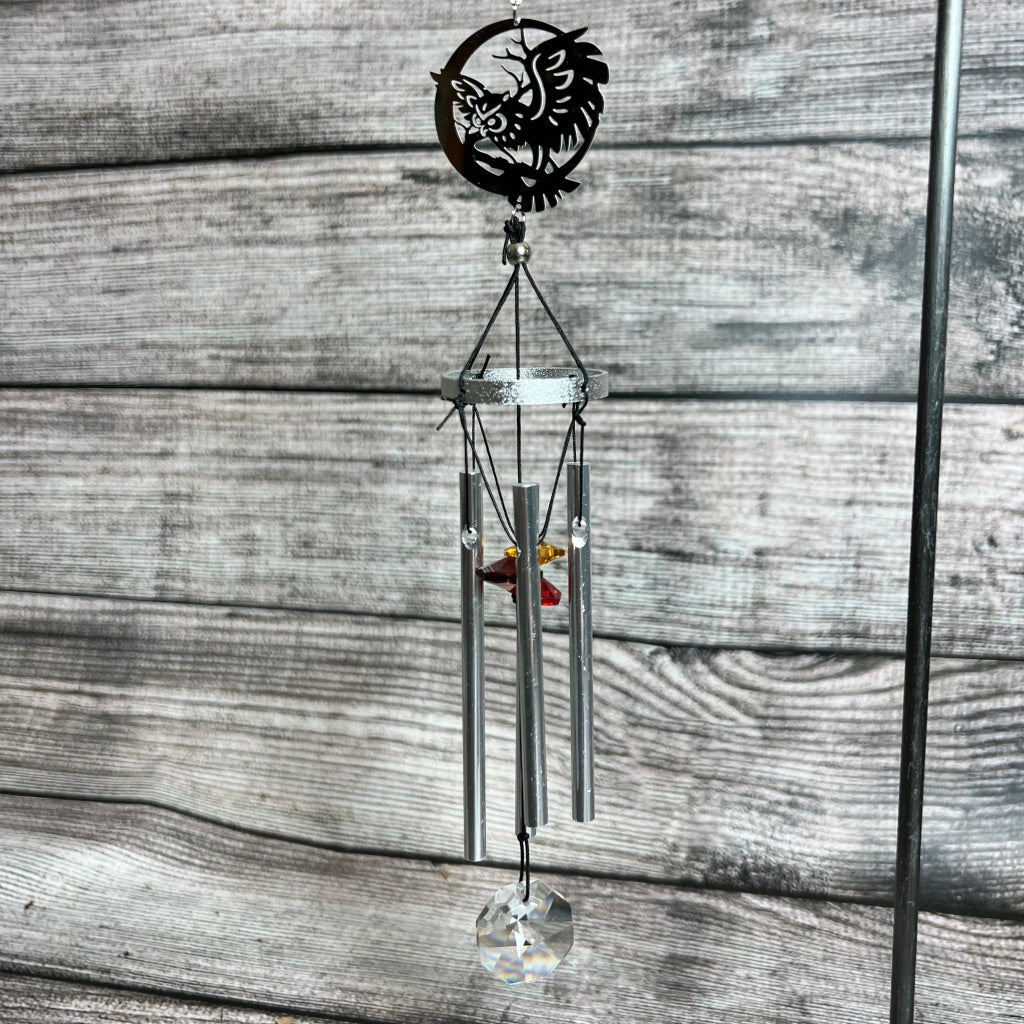 A lovely Sun Chimes featuring an intricate Hamsa hand design at the top. Below it, four slender silver tubes hang, along with a red crystal and a clear prism at the bottom to catch sunlight reflection. The background consists of horizontally oriented wooden planks.
