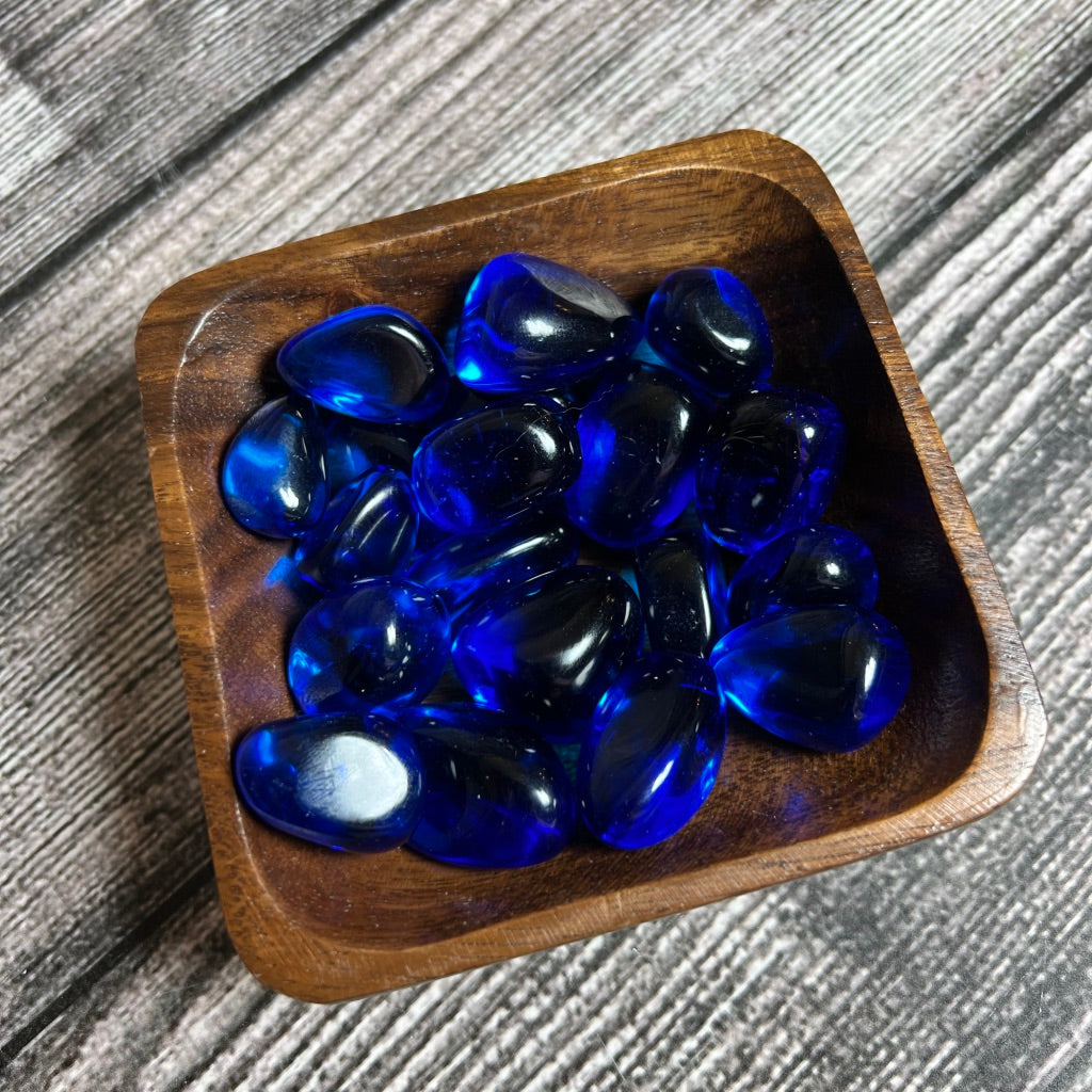 A small wooden bowl filled with polished, Indigo Obsidian rests on a textured wooden surface. The stones, smooth and shiny, reflect light, emphasizing their vibrant color and glossy finish – akin to tiny portals to the spiritual plane.