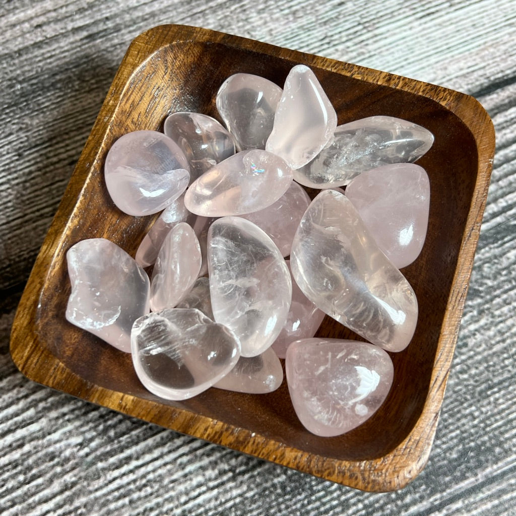 A wooden bowl filled with polished Rose Quartz [high grade] stones sits on a textured wooden surface. The stones vary in size and have a translucent, pink hue, known for their connection to the heart chakra. The bowl's natural wood grain complements the smooth, glossy appearance of the quartz.