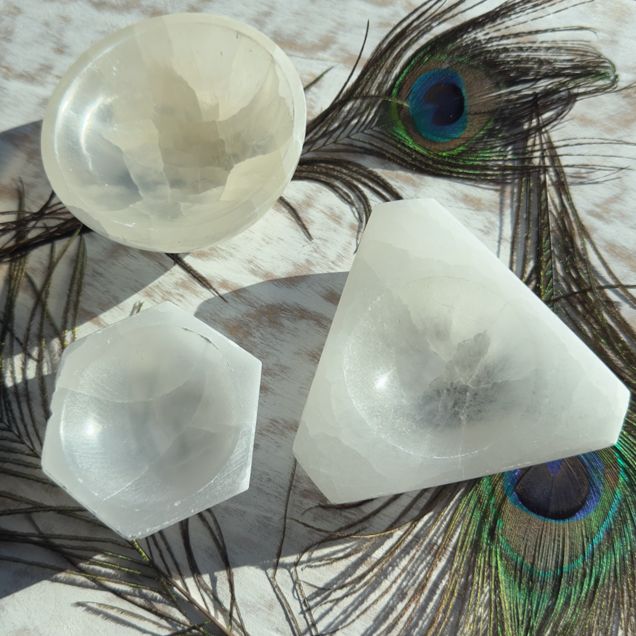 A group of white Selenite Bowls of various shapes (hexagonal, triangular, and round), ideal for meditation and spiritual work, are placed on a light wooden surface. Peacock feathers with vibrant green and blue eye patterns are scattered around them.