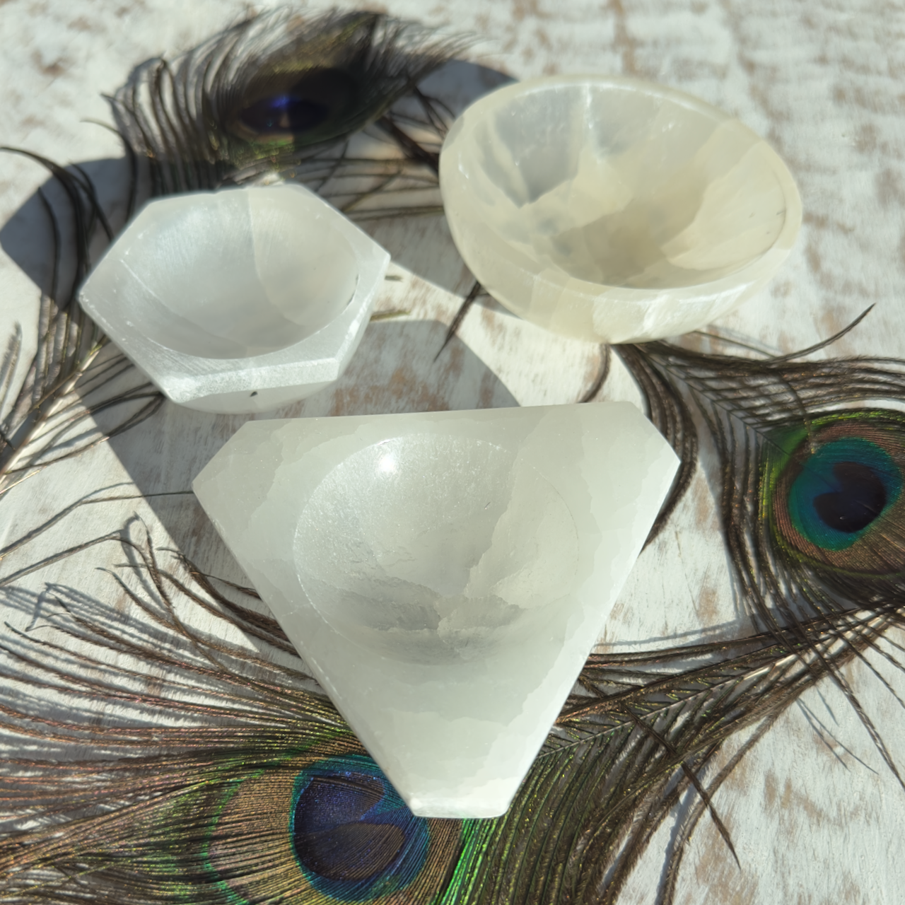 A group of white Selenite Bowls of various shapes (hexagonal, triangular, and round), ideal for meditation and spiritual work, are placed on a light wooden surface. Peacock feathers with vibrant green and blue eye patterns are scattered around them.