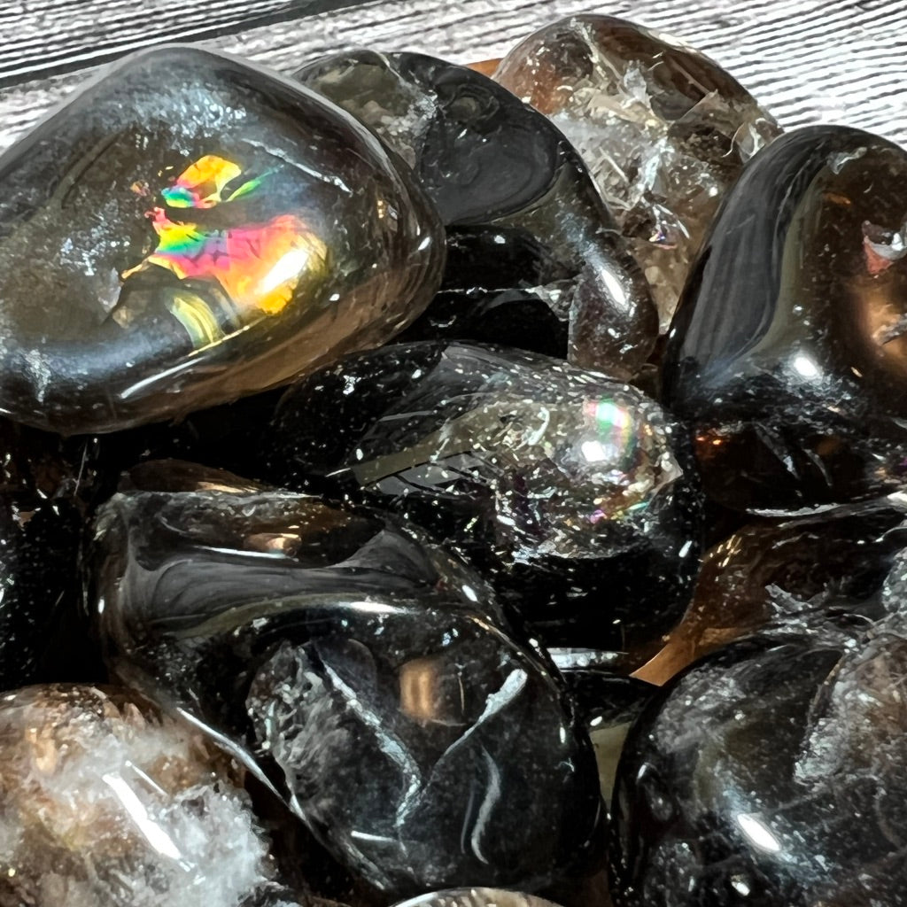 A wooden bowl filled with polished Smoky Quartz [high-grade] stones and pieces of smokey quartz is displayed on a textured, grey wooden surface. Some stones exhibit iridescent patterns, adding a colorful touch and calming energy to the otherwise dark collection.