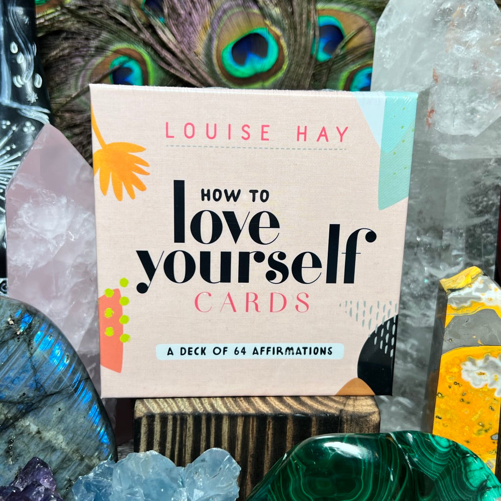 A photo of a card deck  titled 'How to Love Yourself Cards' by Louise Hay, surrounded by decorative crystal figures and stones.