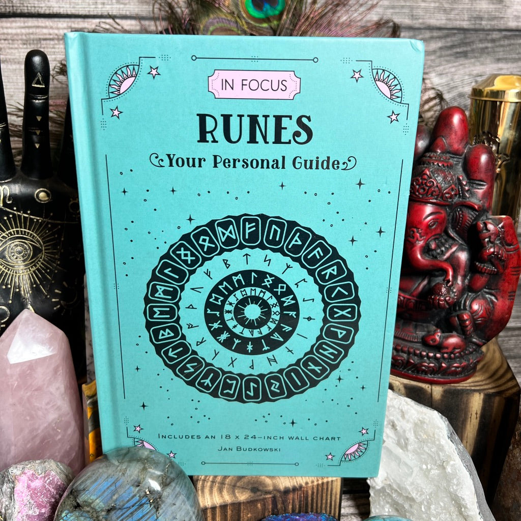 A teal book titled "In Focus Runes: Your Personal Guide" by Jan Budkowski is surrounded by various crystals and statues. The cover features runic symbols in a circular design inspired by Norse mythology. The background includes a wooden surface and various decorative items.