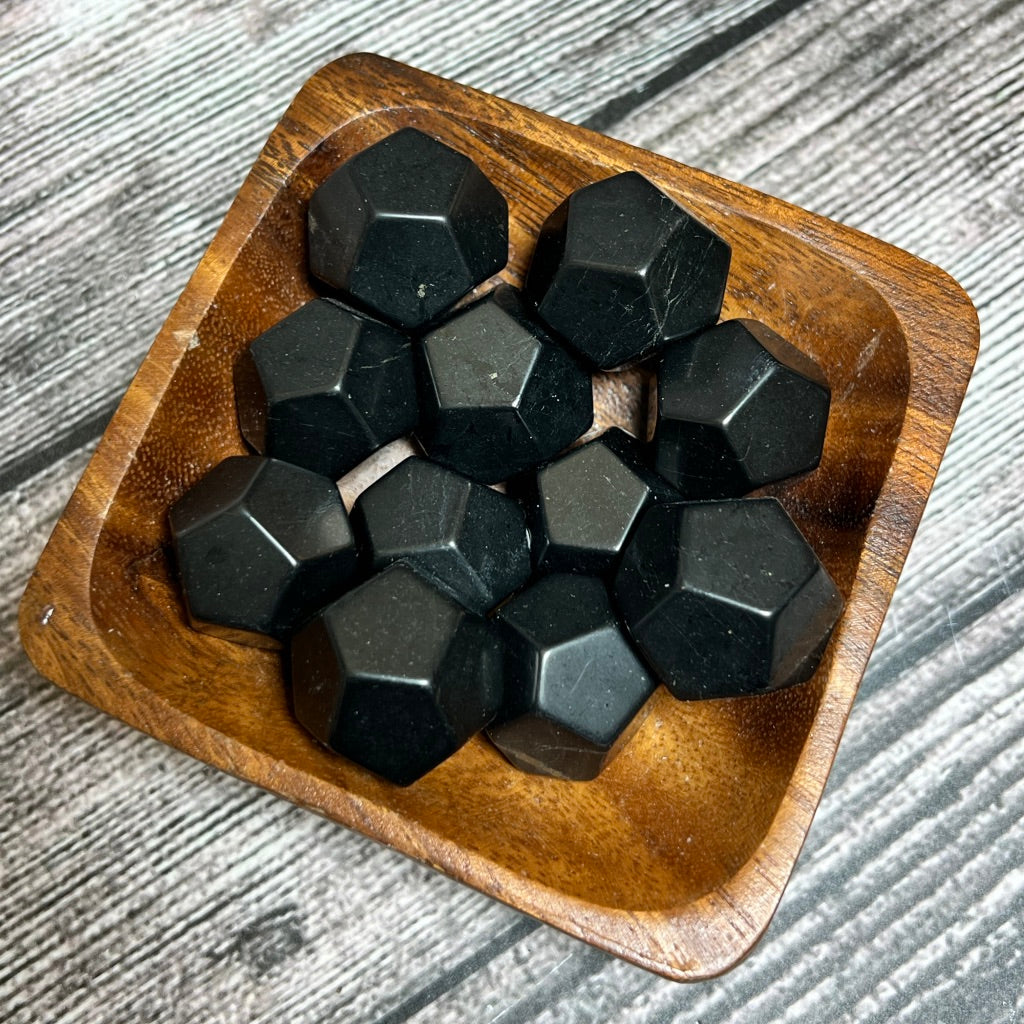 A small wooden bowl holds a collection of Shungite dodecahedrons, reminiscent of Shungite's grounding properties. The bowl rests on a surface with gray, wood-like texture. The dodecahedrons are neatly arranged, showing their faceted surfaces.
