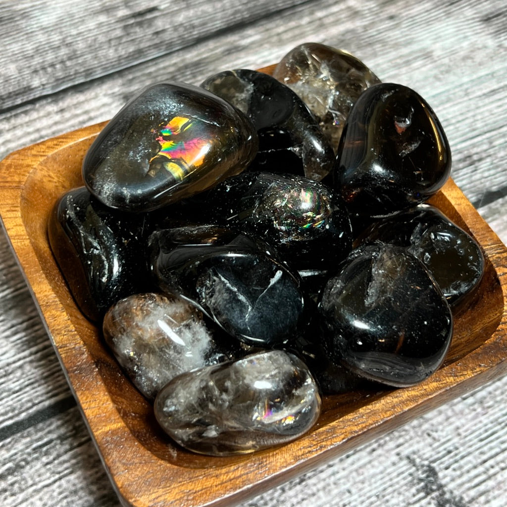A wooden bowl filled with polished Smoky Quartz [high-grade] stones and pieces of smokey quartz is displayed on a textured, grey wooden surface. Some stones exhibit iridescent patterns, adding a colorful touch and calming energy to the otherwise dark collection.
