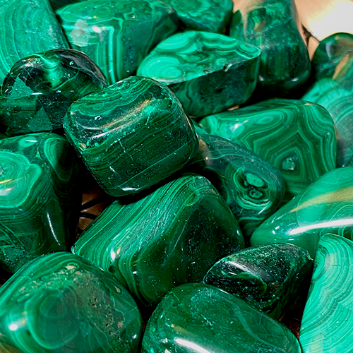 A copper bowl on a wooden surface is filled with polished, green Malachite, known as the "stone of transformation," featuring banded patterns of varying shades of green. The bowl has a geometric design etched on its inner surface, visible around the stones.