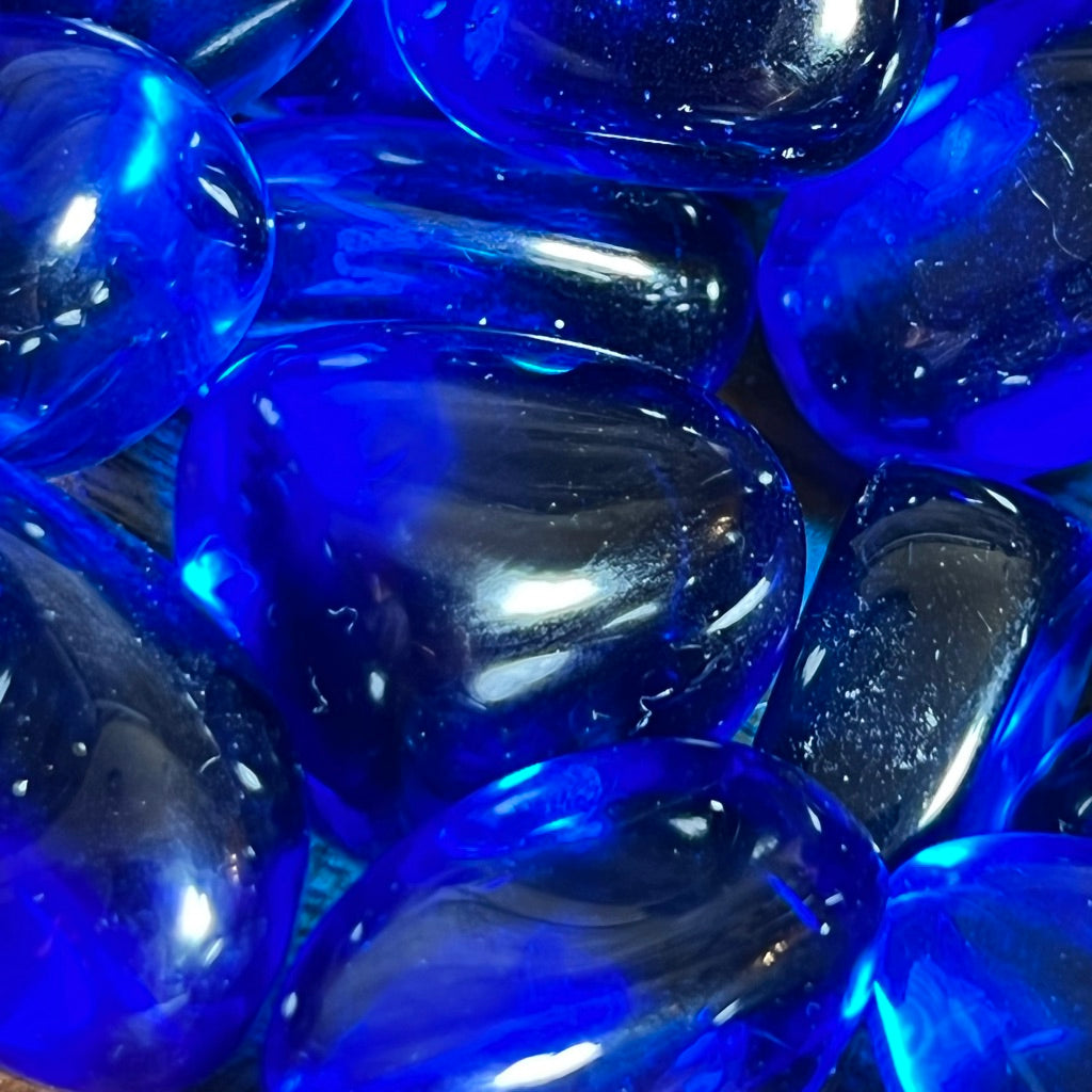 A small wooden bowl filled with polished, Indigo Obsidian rests on a textured wooden surface. The stones, smooth and shiny, reflect light, emphasizing their vibrant color and glossy finish – akin to tiny portals to the spiritual plane.